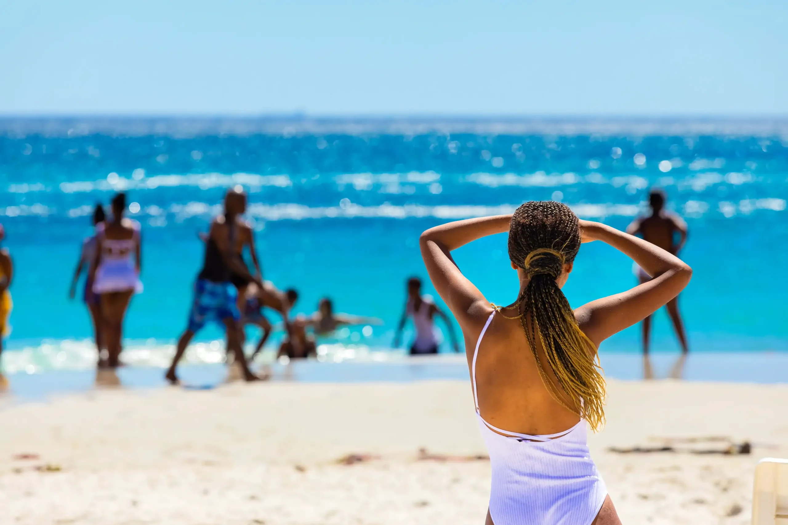 A woman facing turquoise water on a sunny sandy beach, with other beachgoers near the shoreline.