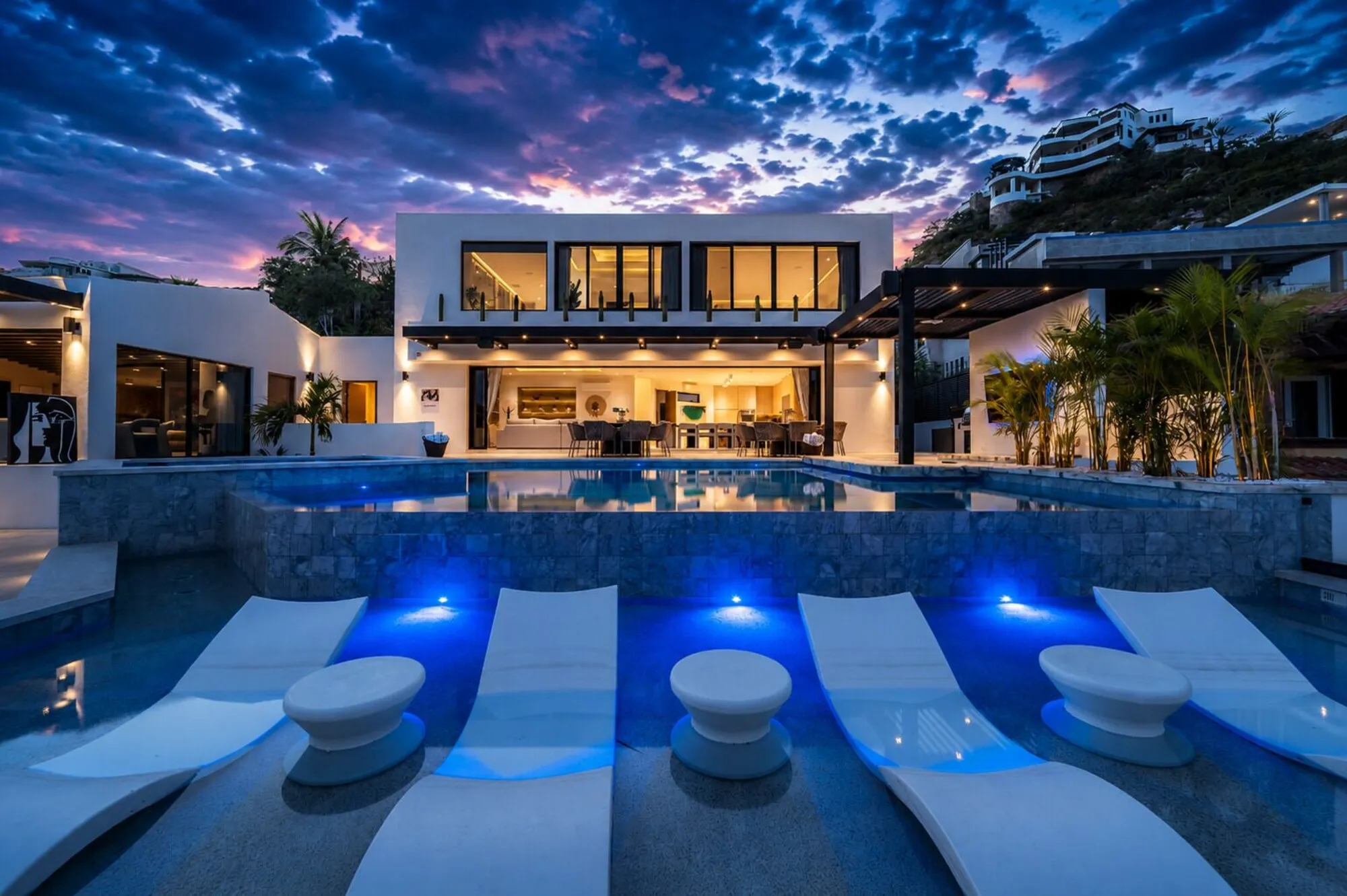 A modern white villa at dusk with illuminated interiors, a blue-lit swimming pool and white in-water loungers.