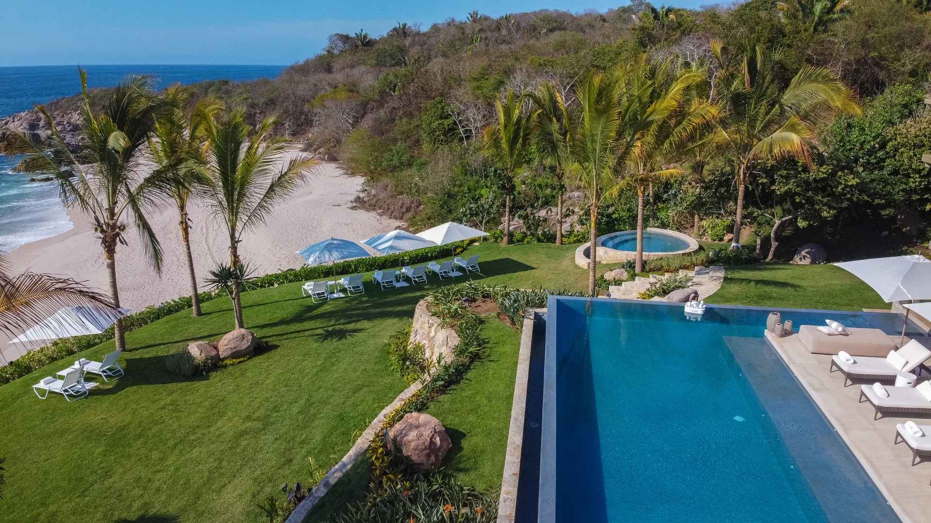 Aerial view of a small sand beach in a rocky cove on the Punta Mita peninsula, framed by palms, with a lawn and an infinity pool on the bluff above it.