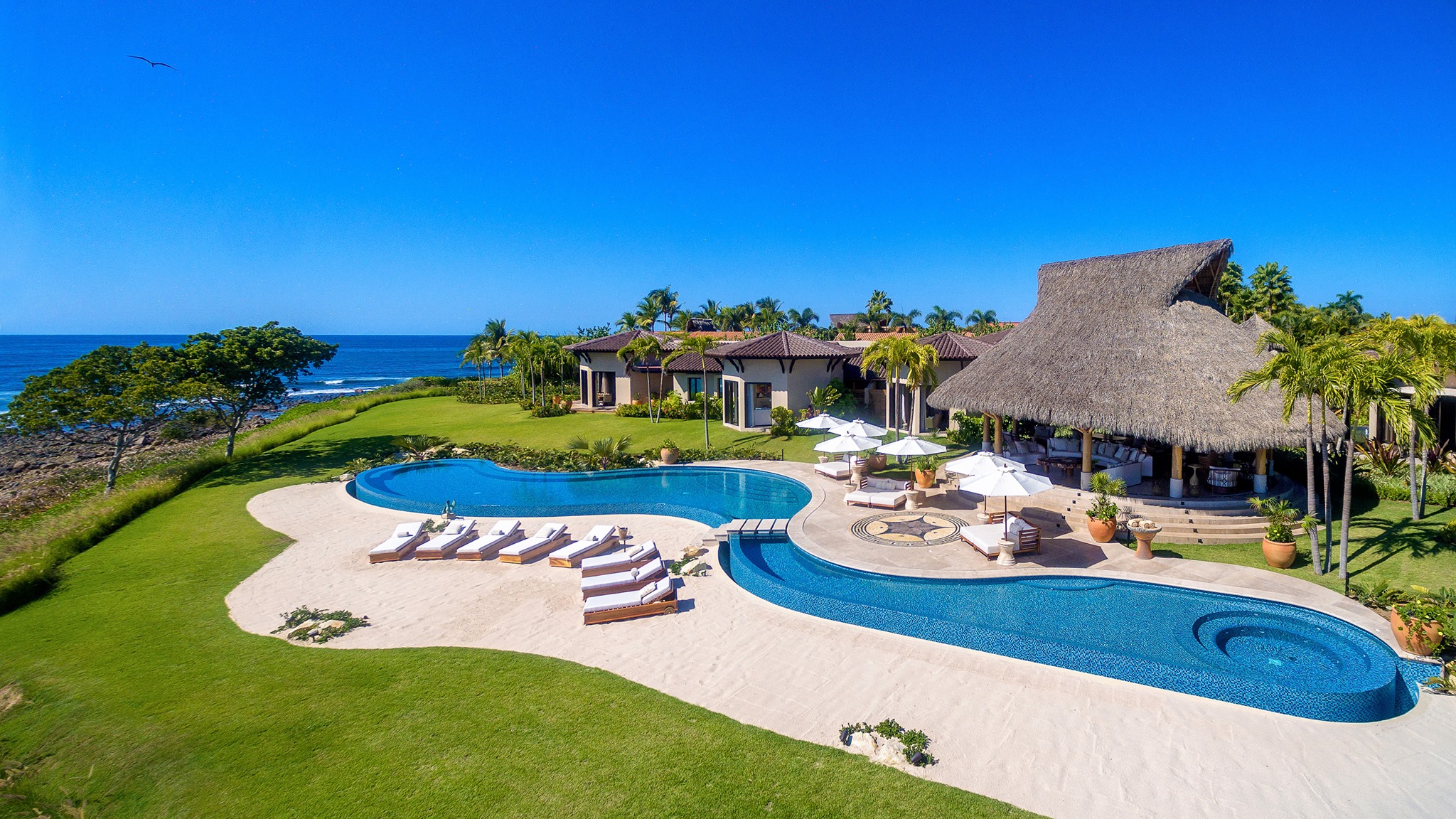 Aerial view of an oceanfront villa on the Punta Mita peninsula: two free-form pools and a sand terrace set in a lawn above a rocky shoreline, with a thatched palapa alongside and the Pacific beyond.