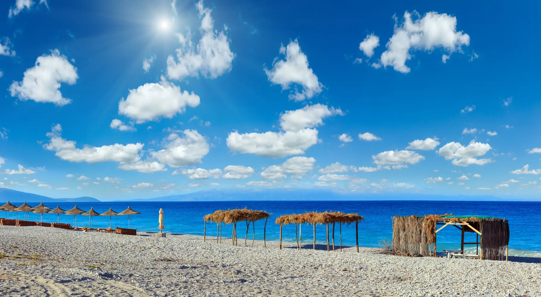 Beach umbrellas beside blue water beneath scattered white clouds.