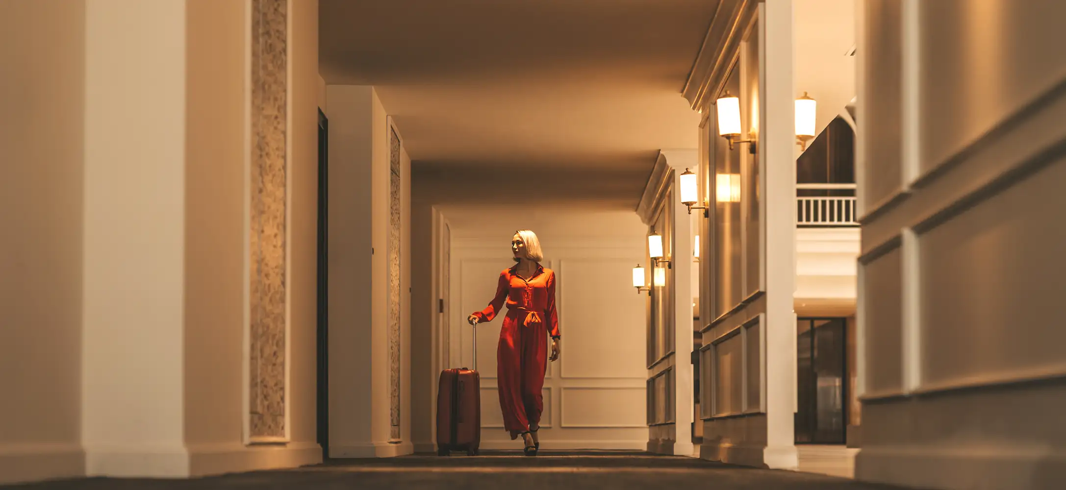 A traveler pulling a suitcase through a warmly lit hotel corridor.