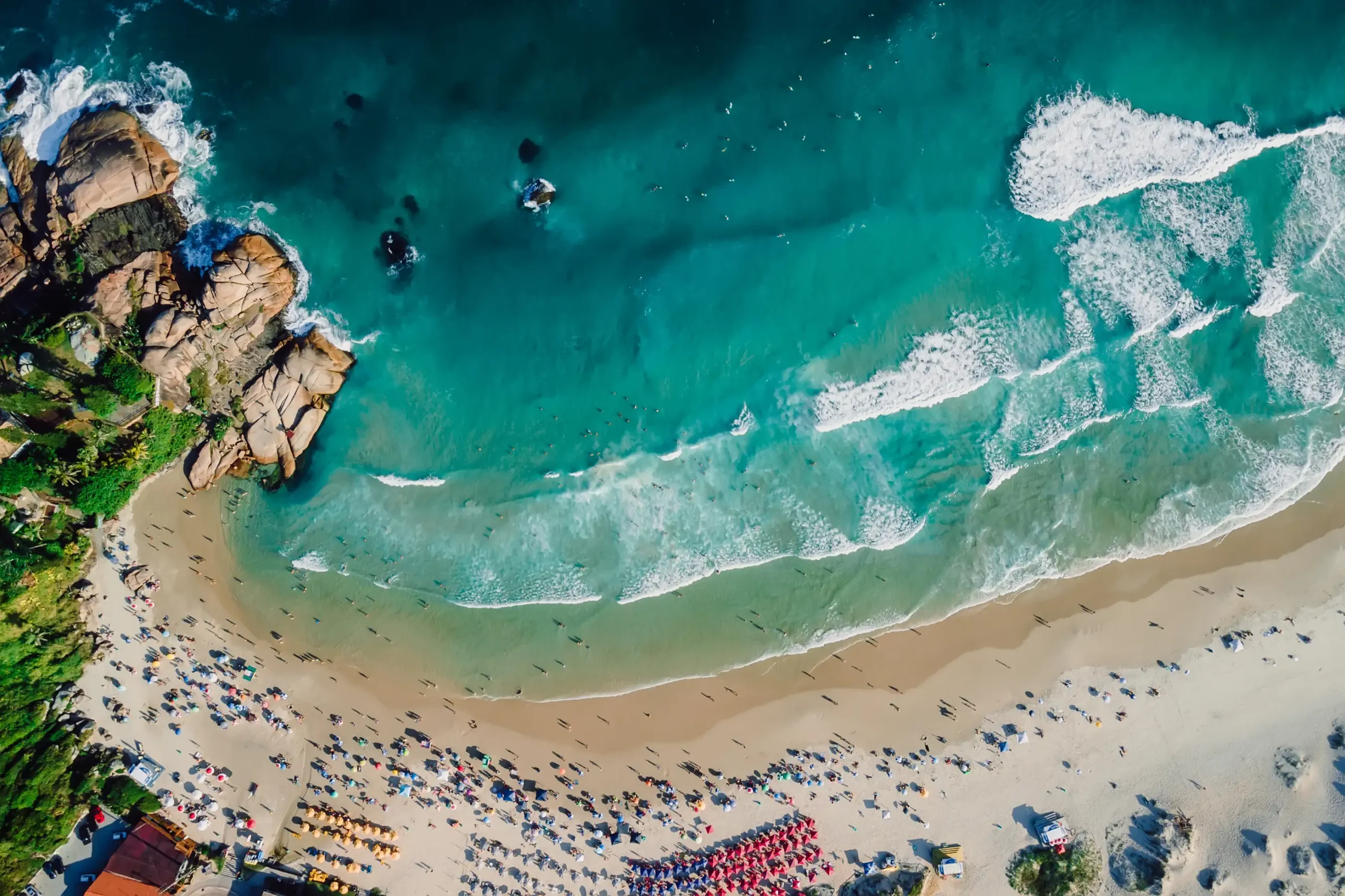 Aerial view of a busy sandy beach beside rocky outcrops and turquoise water with breaking waves.