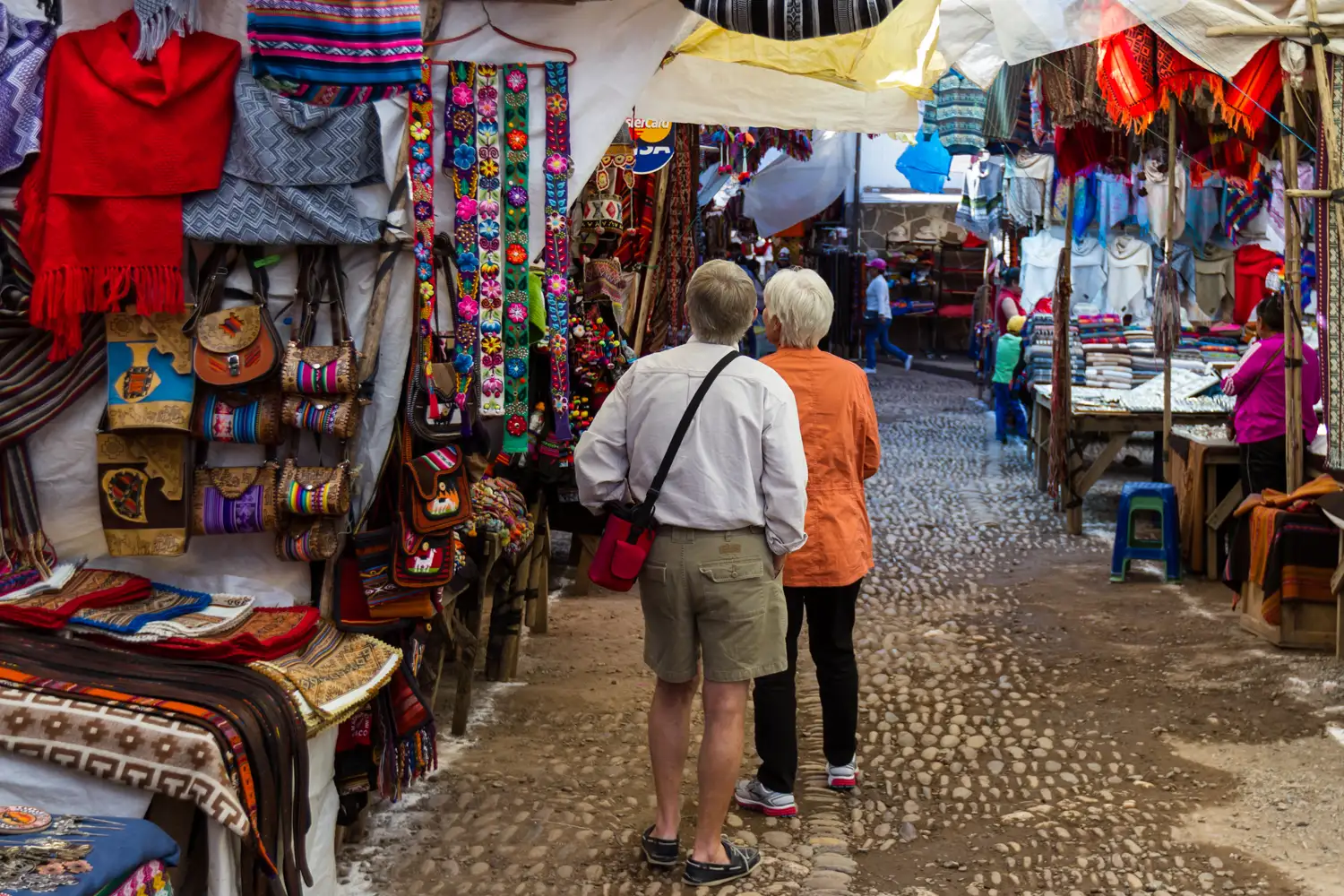 Two visitors walking between colorful textile and souvenir market stalls.