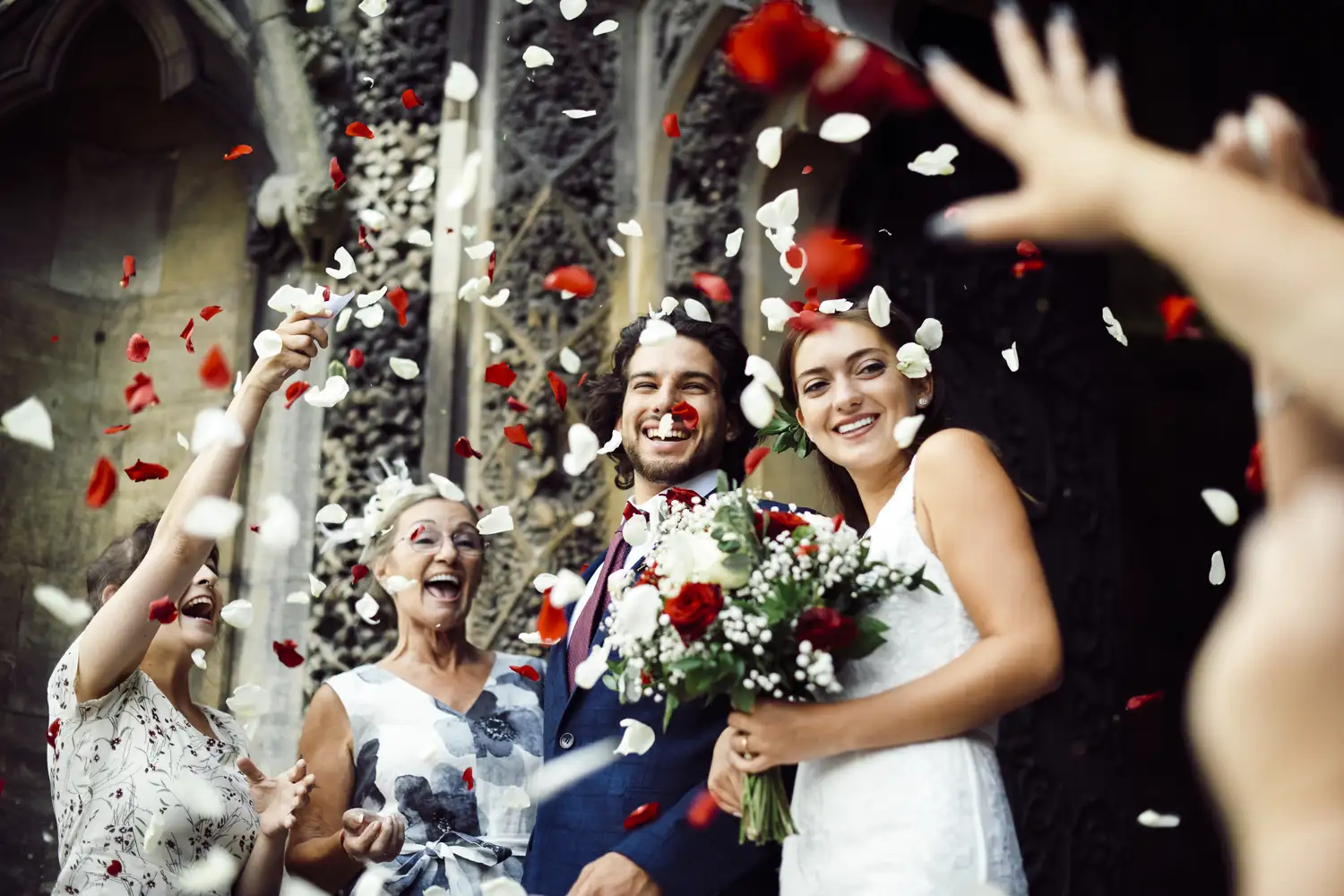 A wedding couple smiling as guests toss red and white flower petals.