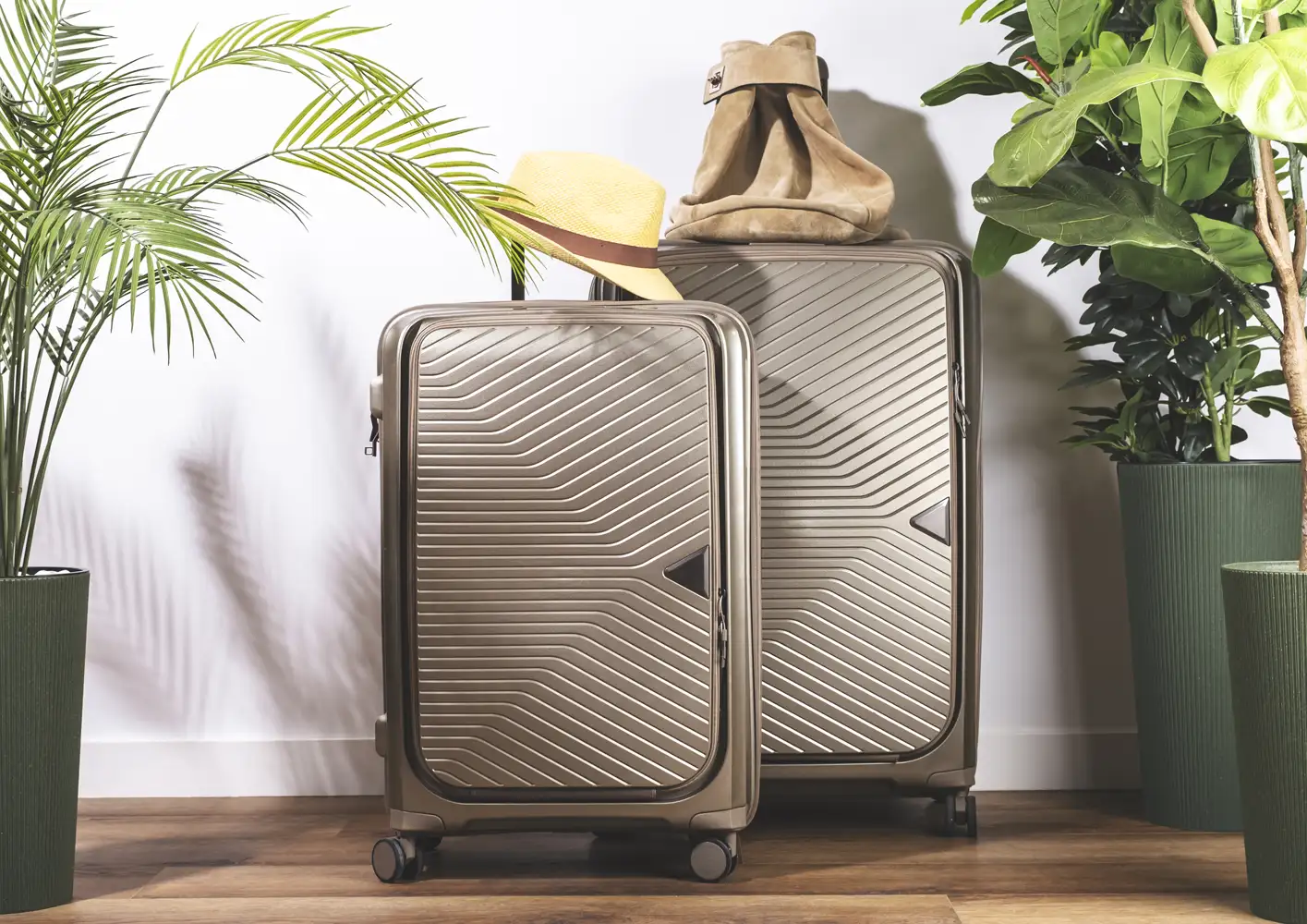 Two wheeled suitcases with a hat and bag beside leafy plants.